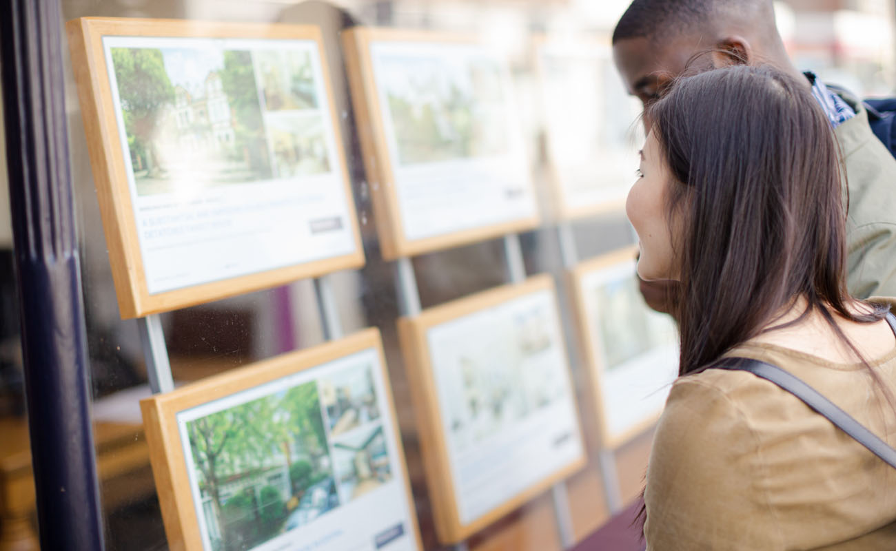 Two people stand outside a real estate agency window, looking at framed property listings with photos and descriptions of homes for sale or rent.